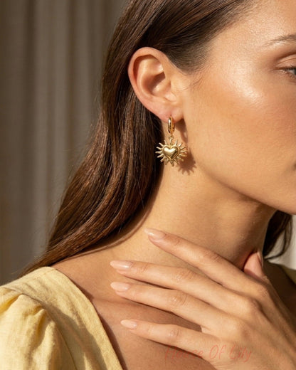 A close-up lifestyle shot of a woman wearing a gold dangle earring with a sunburst and heart design, set against a warm, softly lit background.