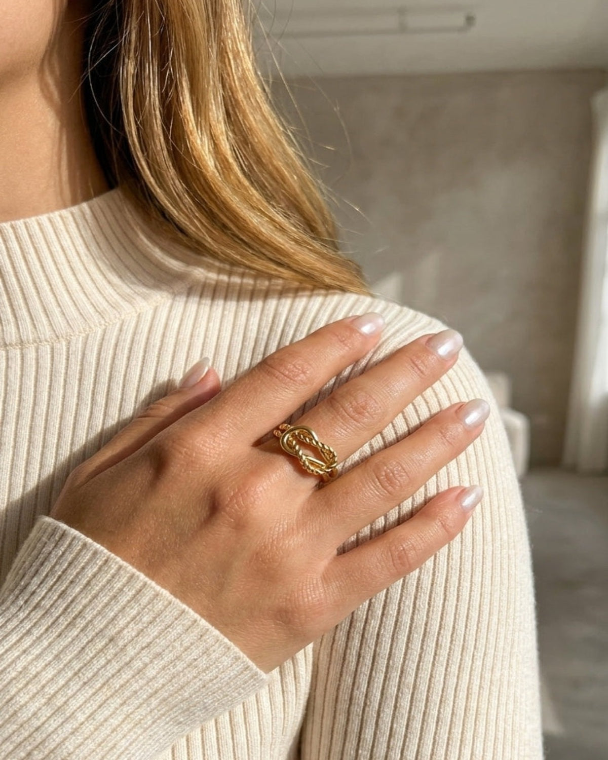 A close-up lifestyle shot of a woman wearing a textured gold ring on her finger, dressed in a cream-colored ribbed sweater. The background is a softly blurred indoor setting.