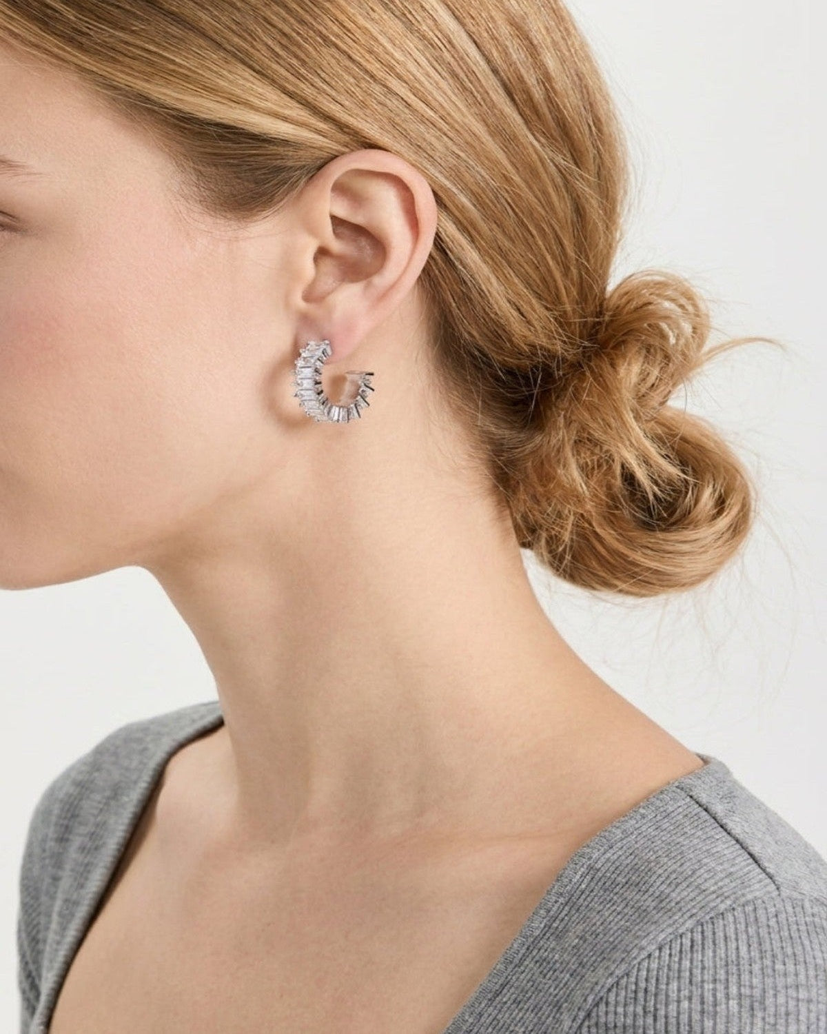 A close-up side profile of a woman with blonde hair in a low bun, wearing a small silver crescent-shaped hoop earring set with clear stones, against a light gray background.