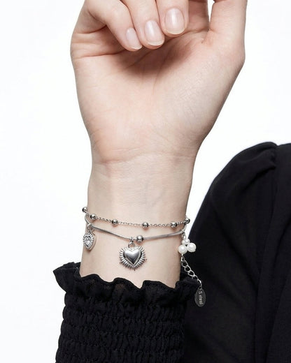 A silver-toned double chain bracelet featuring heart charms and small pearls worn on a wrist.
