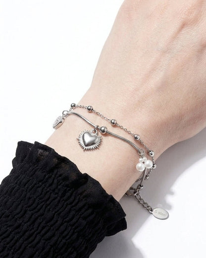 A silver layered chain bracelet with a radiant heart charm and small white pearls on a wrist.