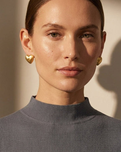 A woman wearing matte gold heart-shaped stud earrings and a grey ribbed top.