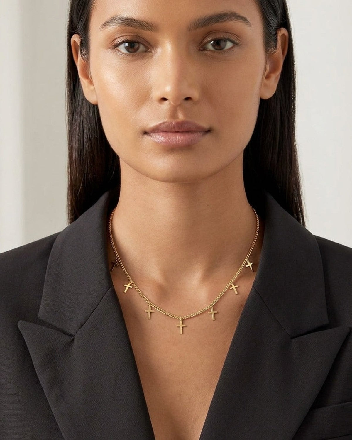 A gold chain necklace with multiple small cross charms worn by a woman in a black blazer.