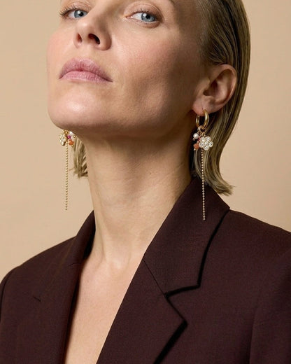 A woman wearing gold beaded flower hoop earrings with hanging chains and a dark brown blazer.