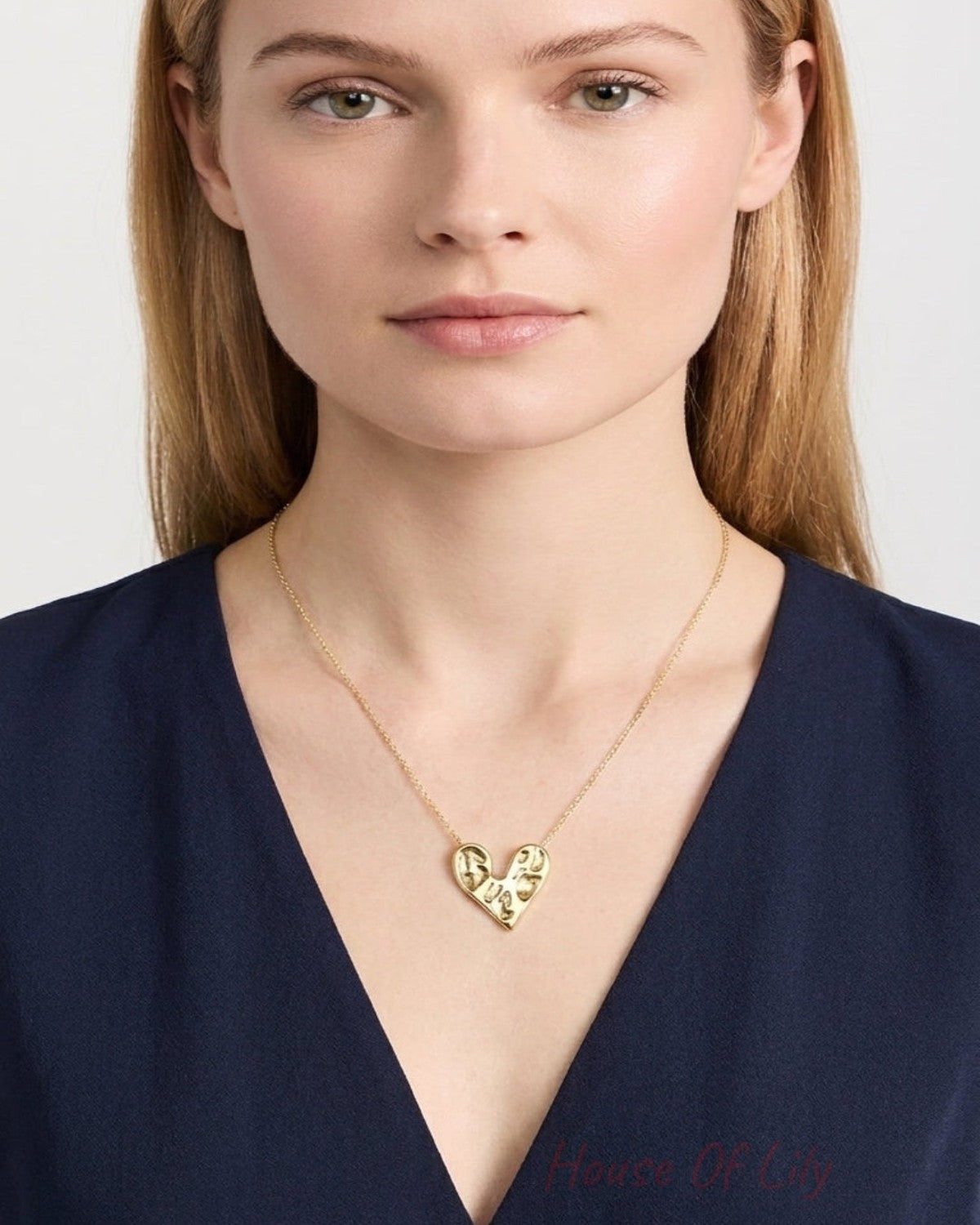 A woman wearing a delicate gold heart pendant necklace with engraved script, paired with a dark navy blue V-neck top, shown against a light gray background.