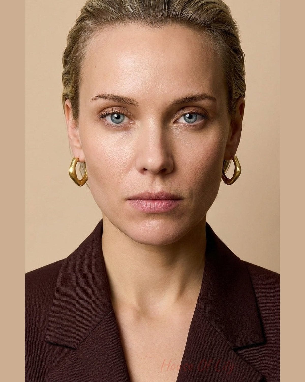 A woman wearing thick, gold-colored rectangular hoop earrings against a neutral beige background.