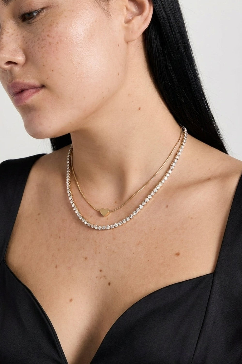 Layered gold necklace featuring a plain chain with a small heart pendant and a diamond-encrusted tennis chain.