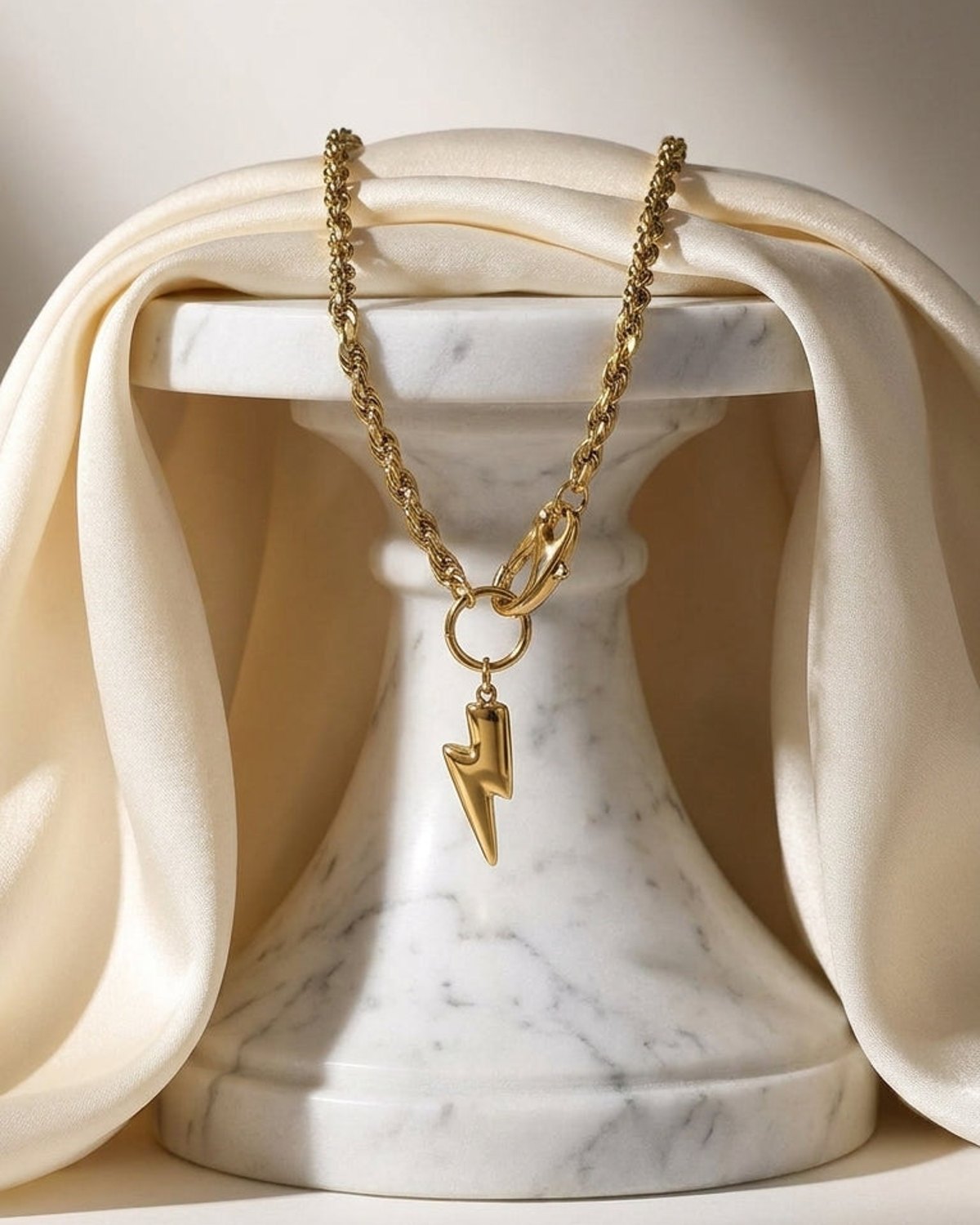 Gold-tone pendant necklace with a textured rope chain and a lightning bolt charm draped over a white marble stand.