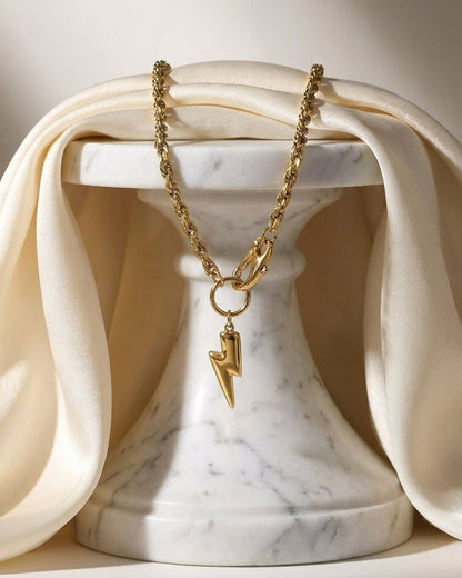 Gold-tone pendant necklace with a textured rope chain and a lightning bolt charm draped over a white marble stand.