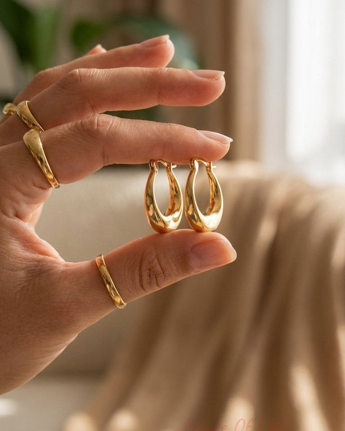 A hand holding a pair of glossy, thick gold hoop earrings with an uneven, melted shape.