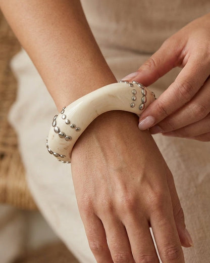 A thick, off-white resin bangle bracelet worn on a wrist, featuring embedded silver-tone metal studs.