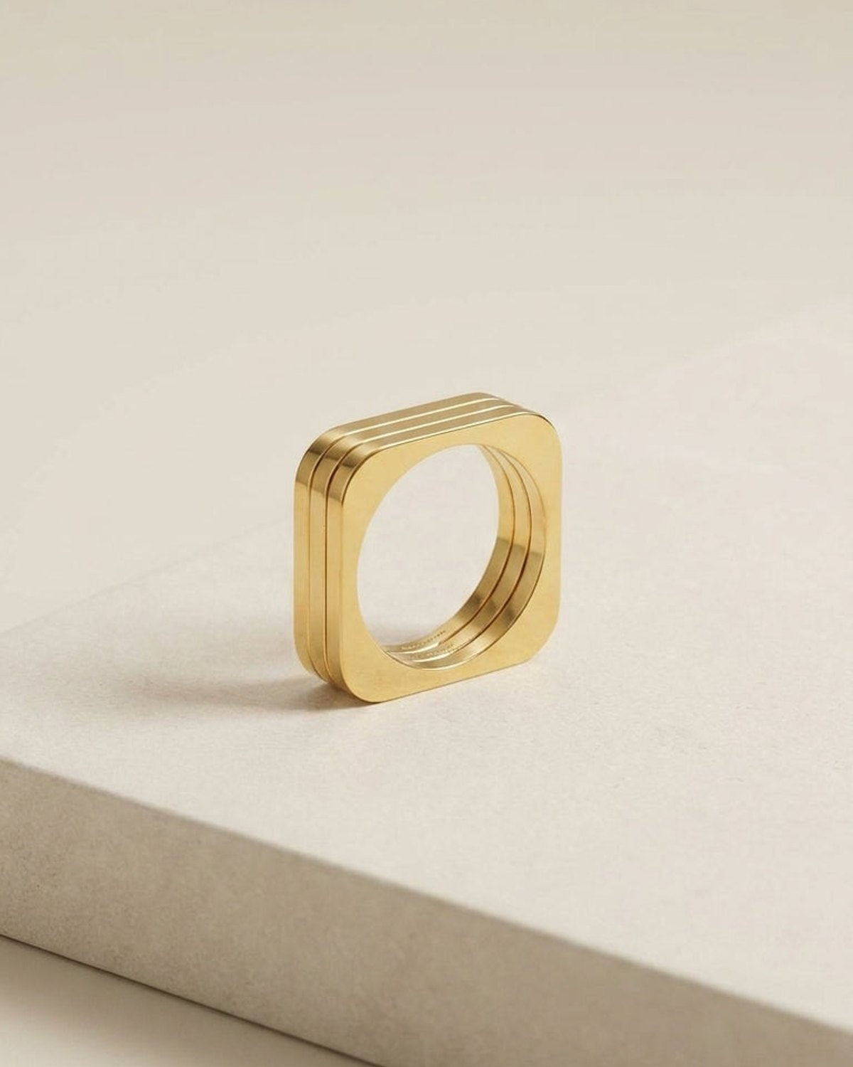 Geometric Edge Gold ring: a gold-colored metal ring with a square shape and rounded corners, comprising three nested bands.