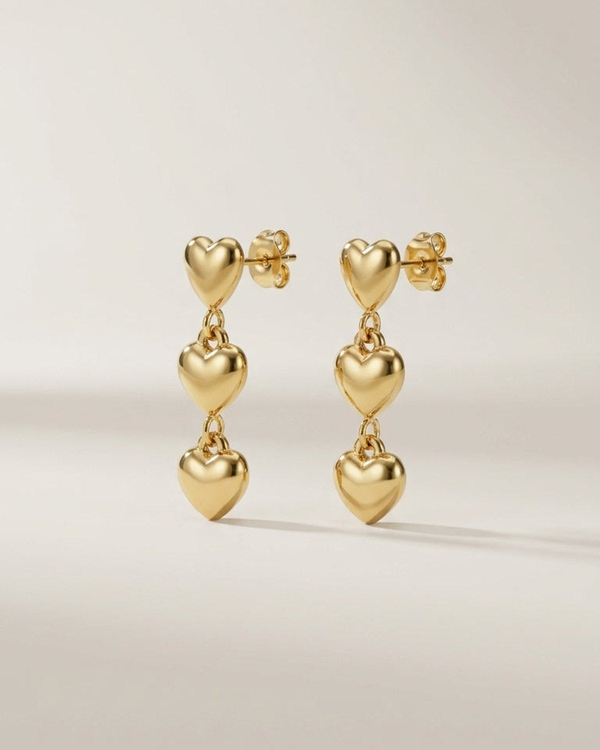 Gold heart dangle earrings, each featuring three puffed hearts linked vertically on post backs.
