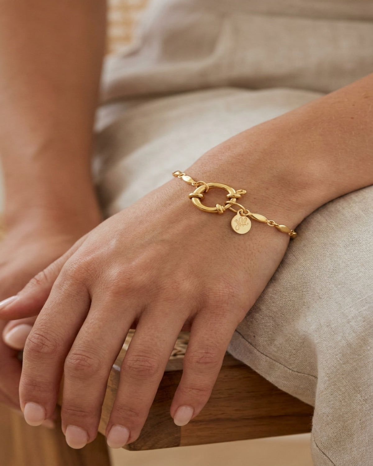 Gold oval-link chain bracelet with a round clasp and small monogram pendant on a wrist.