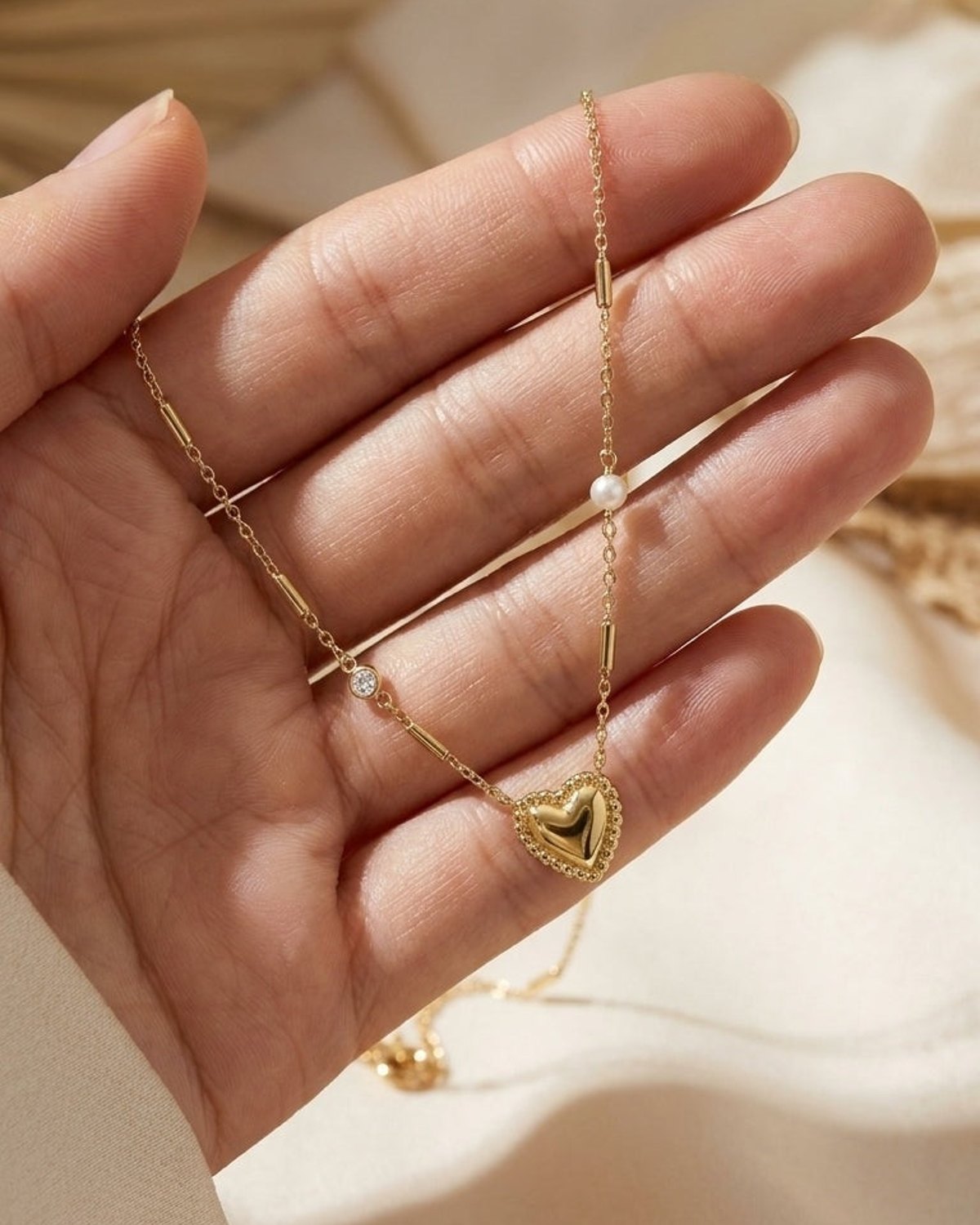 A hand holds a gold heart pendant necklace with interspersed stones and pearls against a neutral background.
