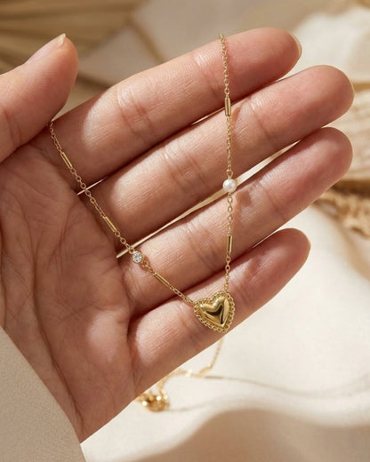 A hand holds a gold heart pendant necklace with interspersed stones and pearls against a neutral background.