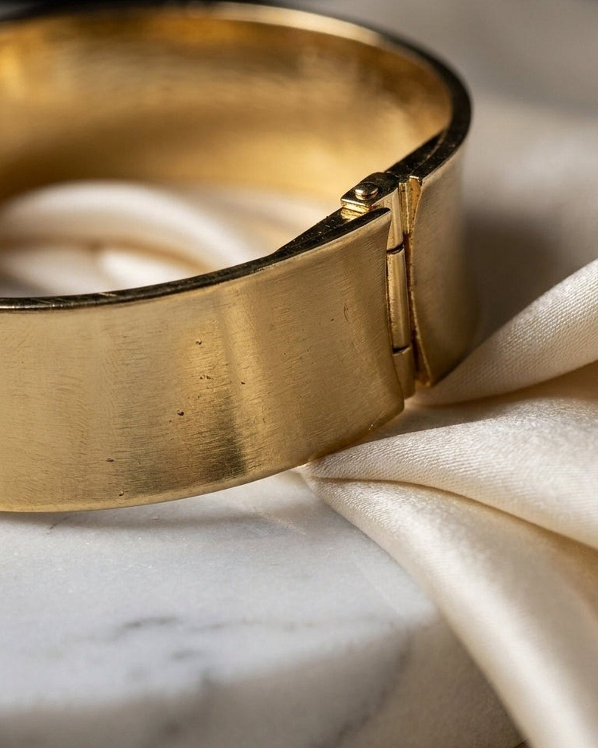 Brushed gold hinged bangle resting on white marble and draped cream silk fabric.
