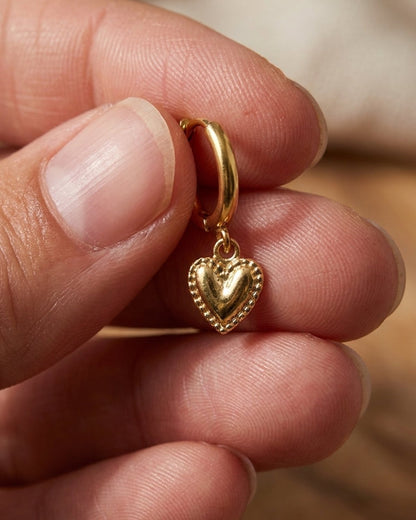 Gold hoop earring with a beaded-edge puffy heart charm held in fingers.