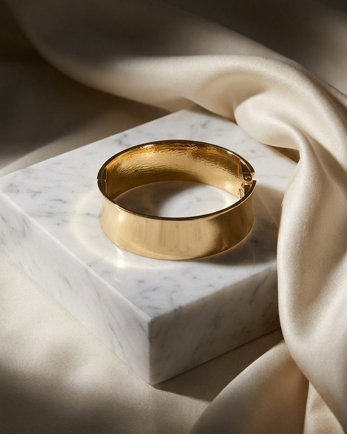 A gold hinged bangle bracelet rests on a white marble block, framed by draped cream fabric.