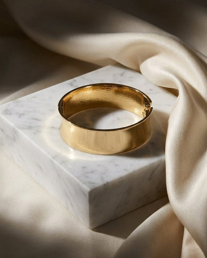 A gold hinged bangle bracelet rests on a white marble block, framed by draped cream fabric.