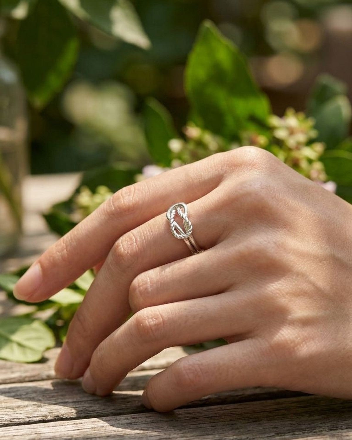 Delicate Silver Twisted Knot ring worn on a model's finger outdoors, with sunlight filtering through leaves and a softly blurred natural background.