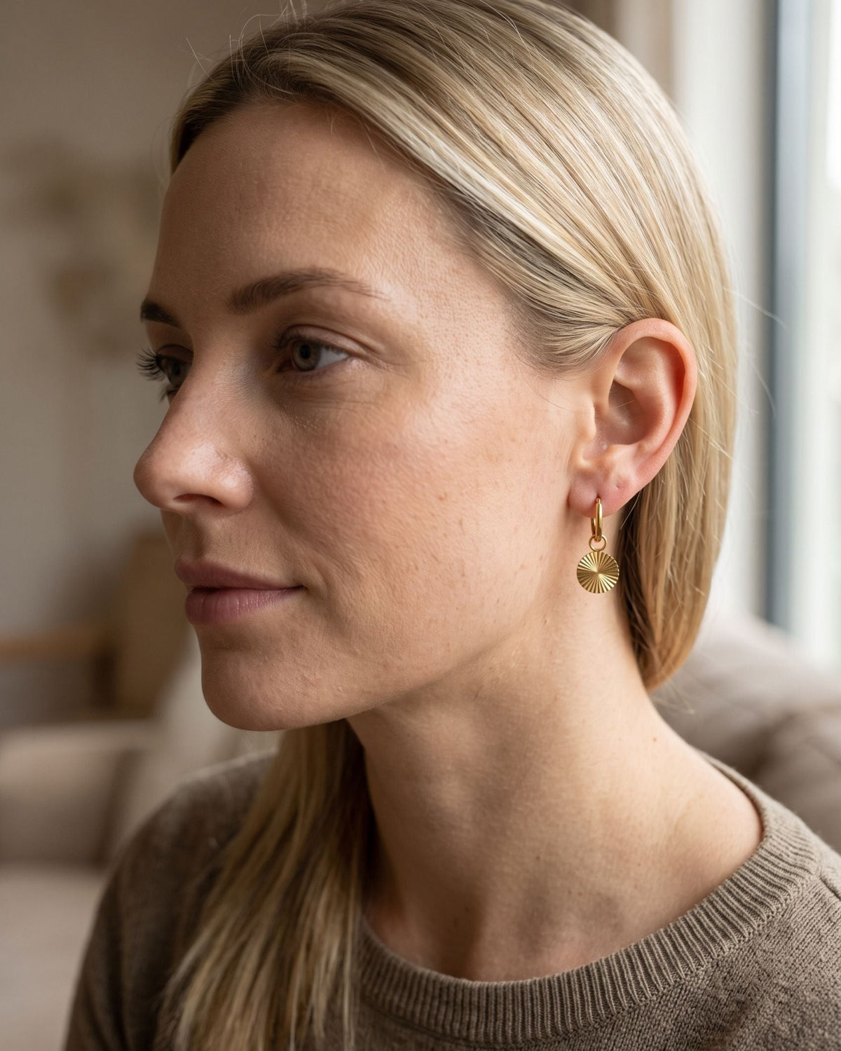 Model wearing minimalist stainless steel latch back hoop earrings with a small textured radiating disc pendant.