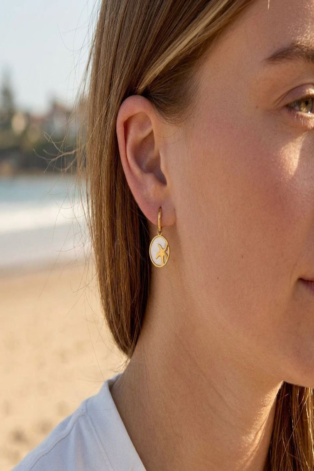 Close-up of starfish glow hoop earrings hanging from a model's earlobes, showcasing details of sea shell elements and stainless steel, with a blurred beach background.