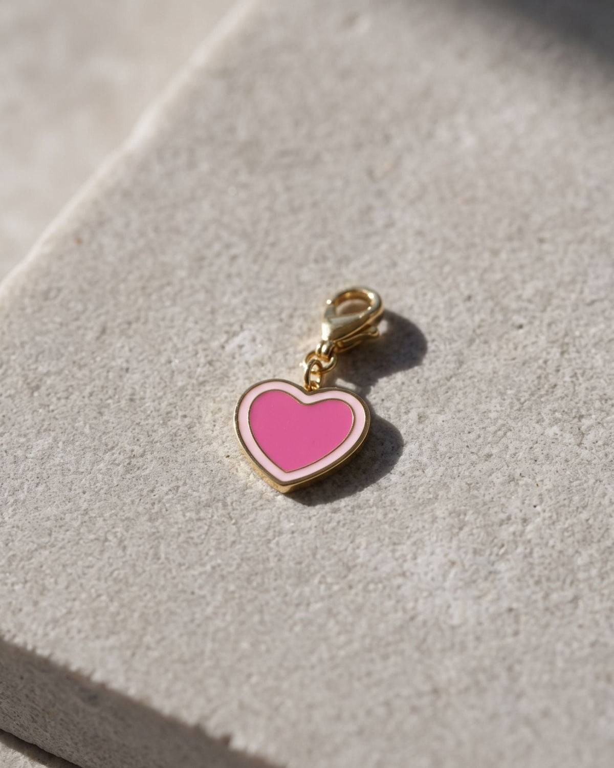 Small heart-shaped enamel charm on a gold-tone lobster clasp resting on a textured white stone surface.