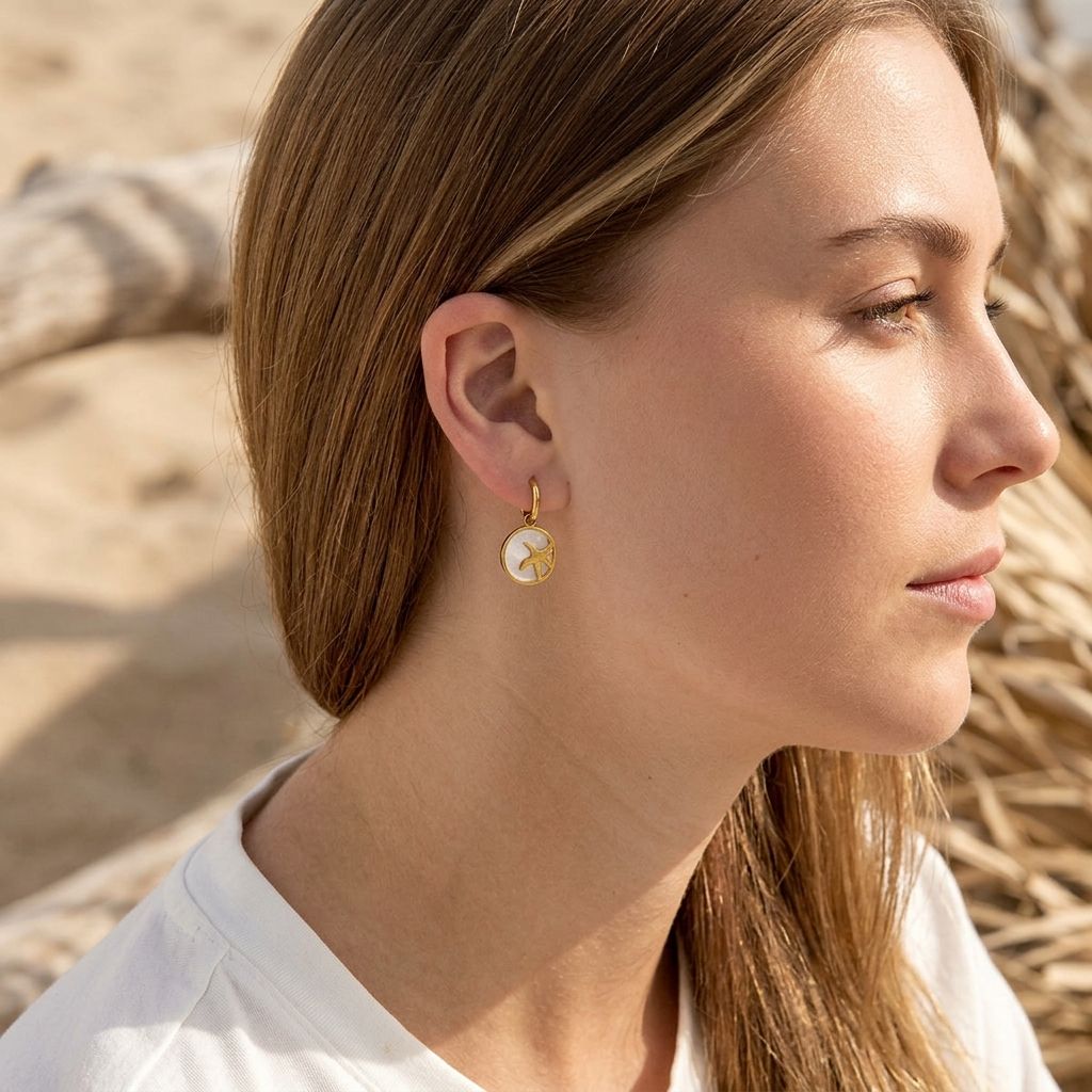 Close-up of starfish glow hoops earrings with shell details worn on a model's ears, highlighted by natural sunlight with beach tones in background.