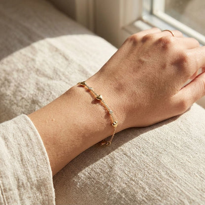 Close-up of dainty sweetheart charm bracelet with rhinestone hearts on a wrist, in natural sunlight on textured fabric.