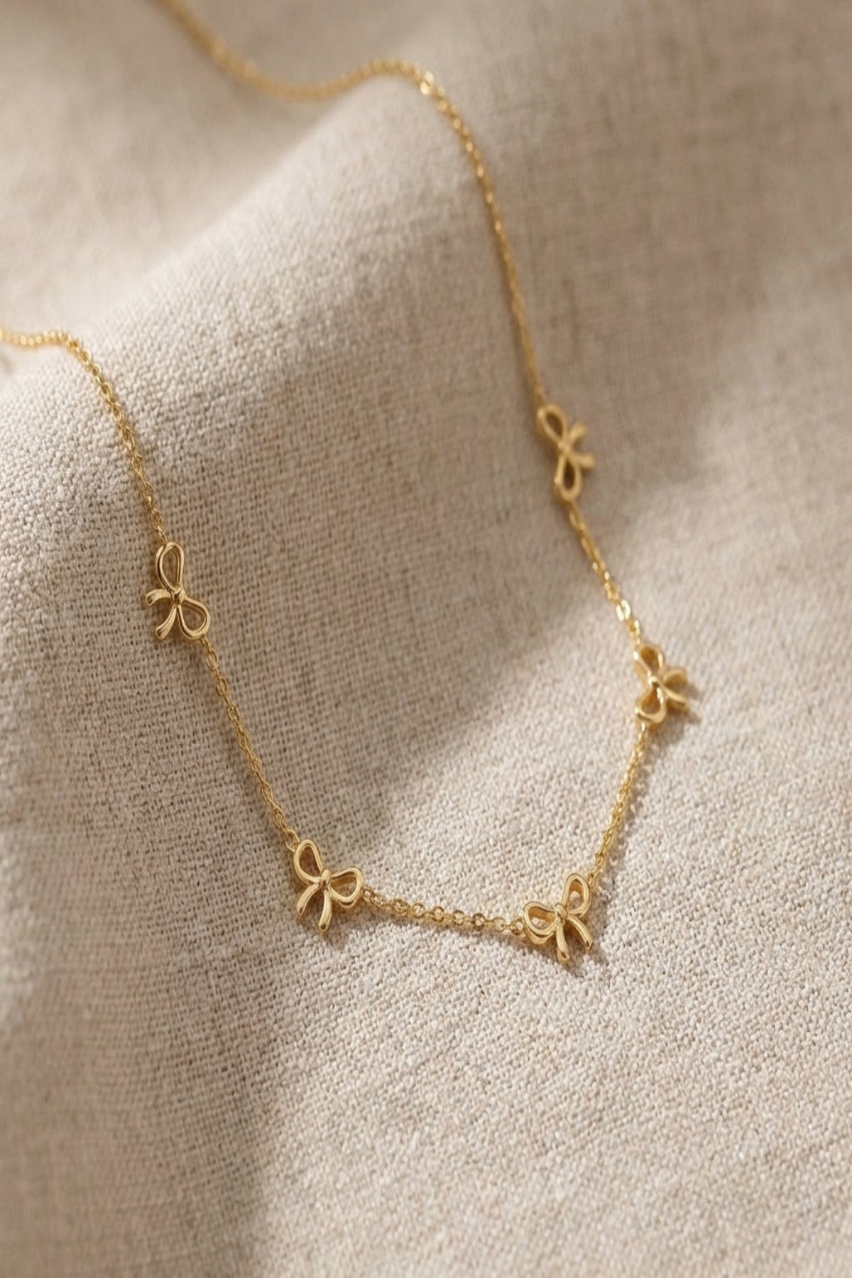Close-up of a delicate stainless steel necklace with five tiny bow charms, displayed on soft fabric with natural lighting to highlight its fine craftsmanship.