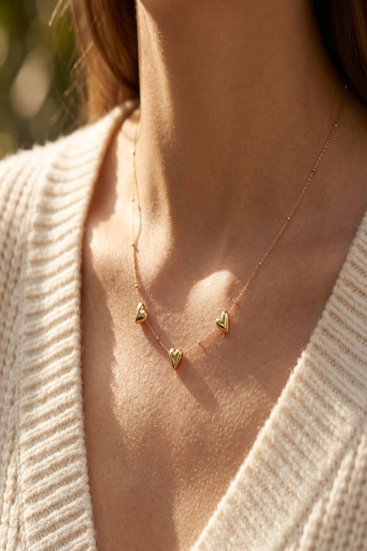 Close-up of a delicate gold necklace with three small heart pendants set against a blurred natural background, highlighting fine craftsmanship and reflective gold surface.