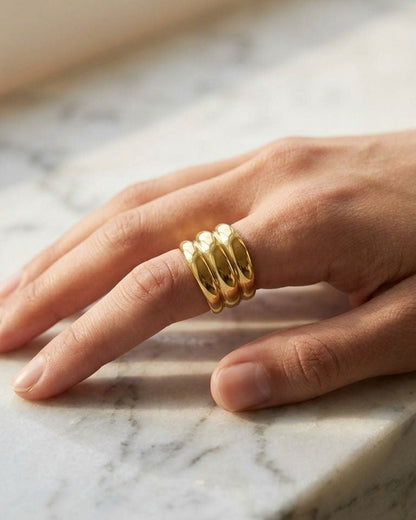 Model wearing Triple Stack Gold ring on finger resting on white marble with warm ambient lighting.
