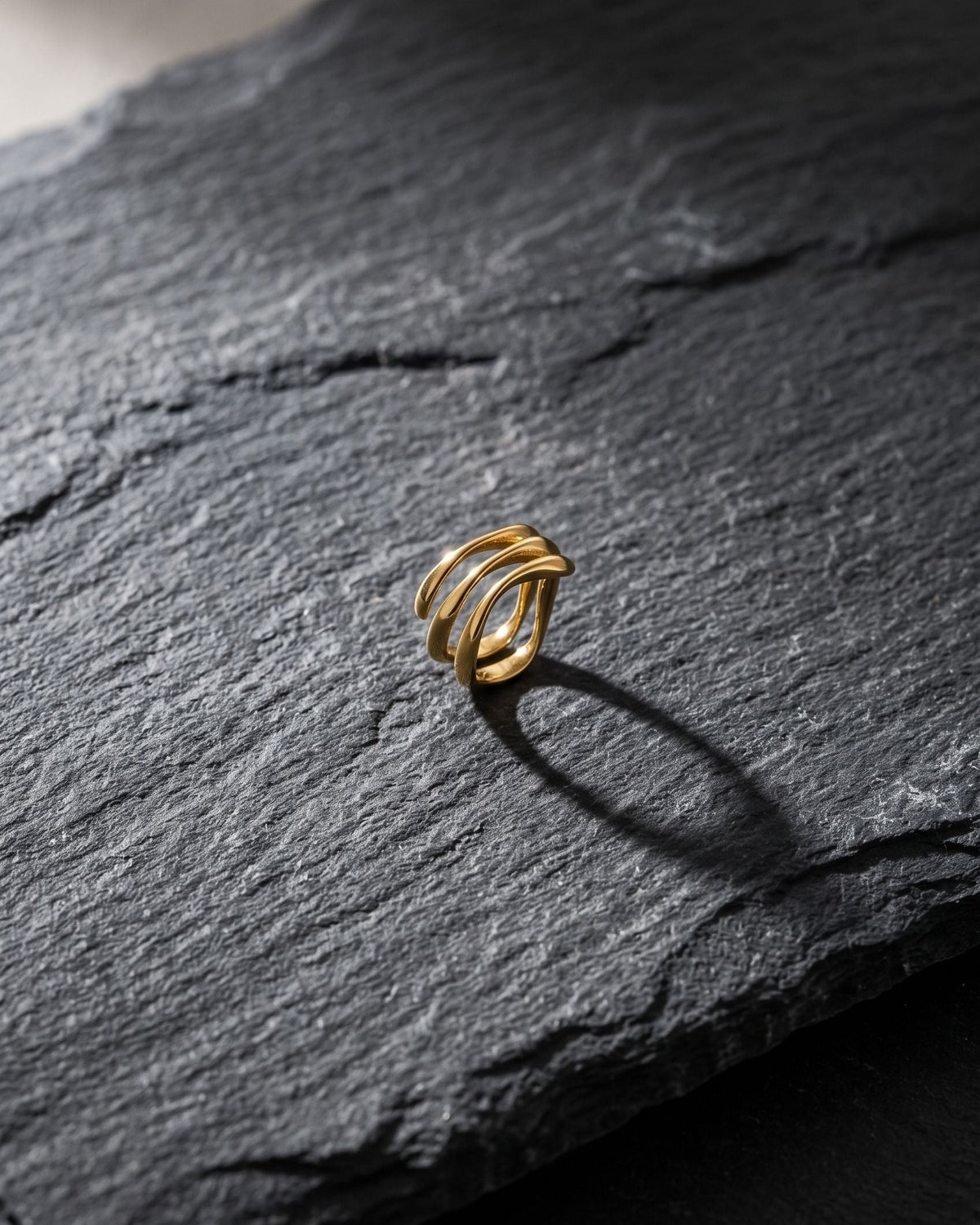 Stainless steel wave stack ring resting on a worn grey slate stone with natural light casting defined shadows.