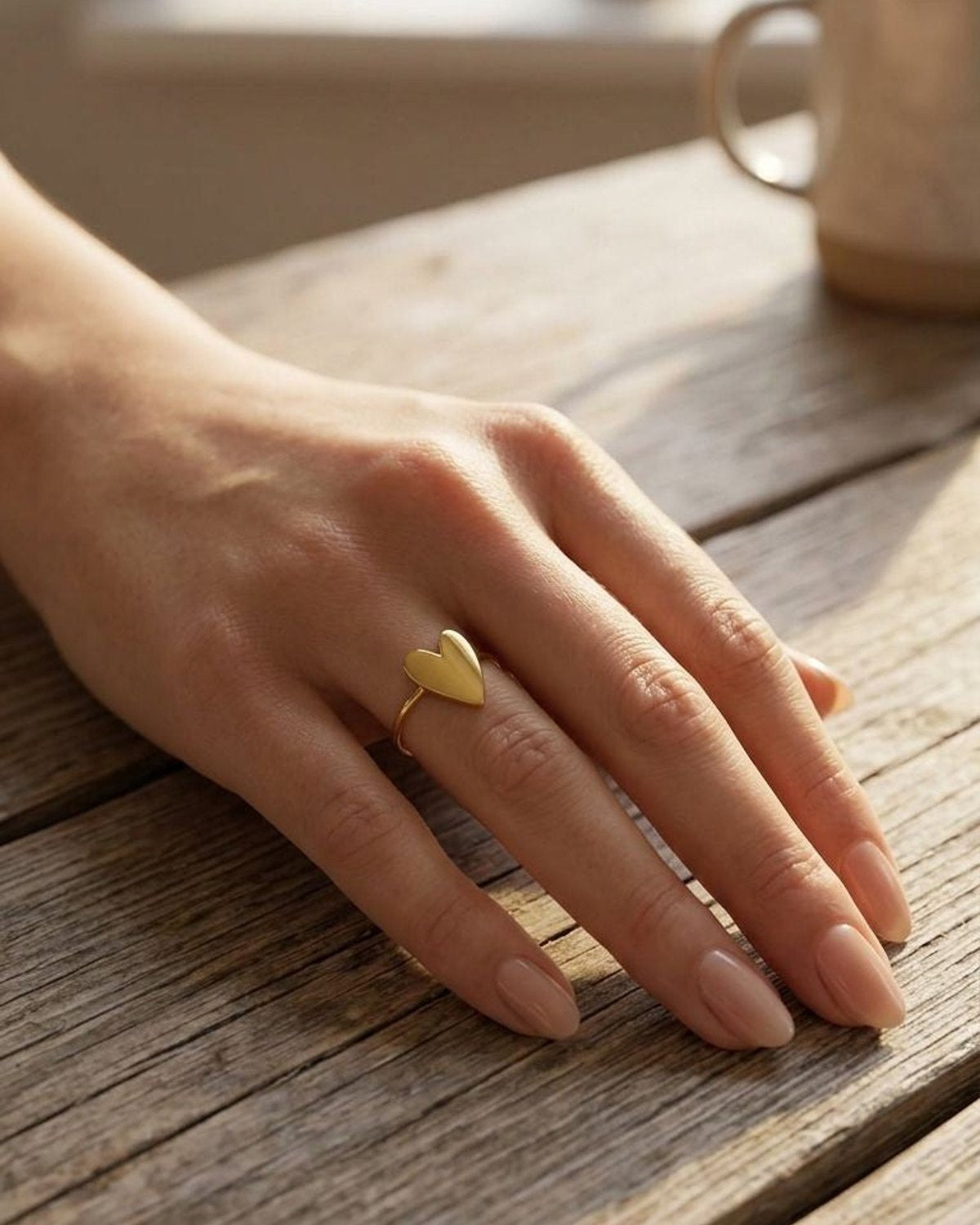 Woman's hand wearing the adjustable Lily Heart ring on a rustic wooden table, showing its dainty, minimalist gold design in natural light.