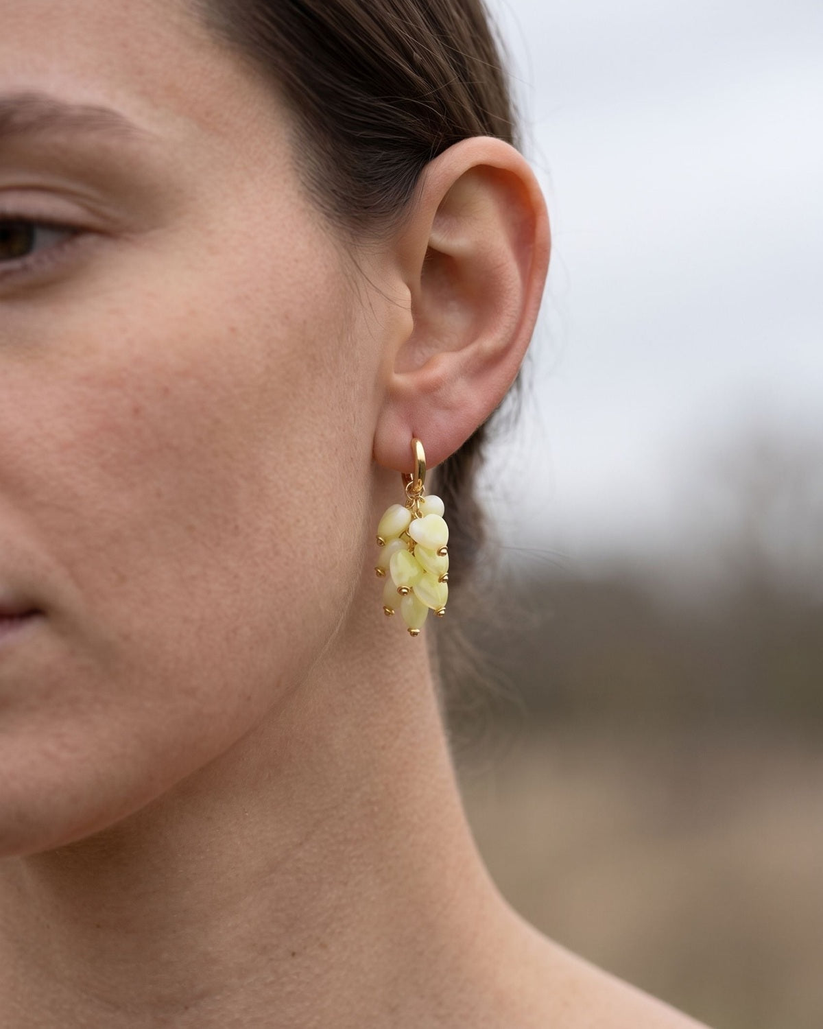 A close-up portrait of a woman wearing a delicate heart-shaped bead earring on her earlobe in an outdoor setting.