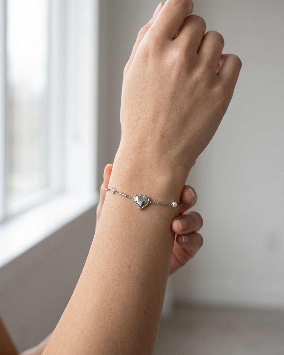 Minimalist stainless steel heart charm bracelet worn on a wrist in soft natural window light.